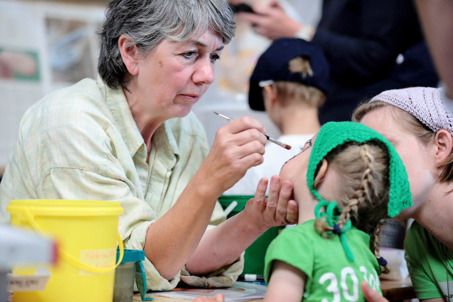 KI generiert: Eine Frau malt das Gesicht eines Kindes an, während weitere Menschen im Hintergrund beschäftigt sind.