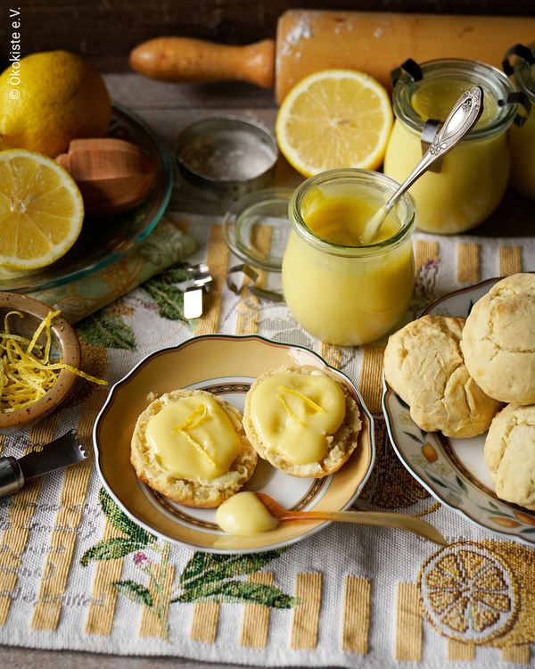 KI generiert: Das Bild zeigt eine kulinarische Szene mit frischem Lemon Curd in einem Glas, umgeben von Zitronen und Gebäck. Im Vordergrund sind zwei Gebäckstücke mit Lemon Curd bestrichen.