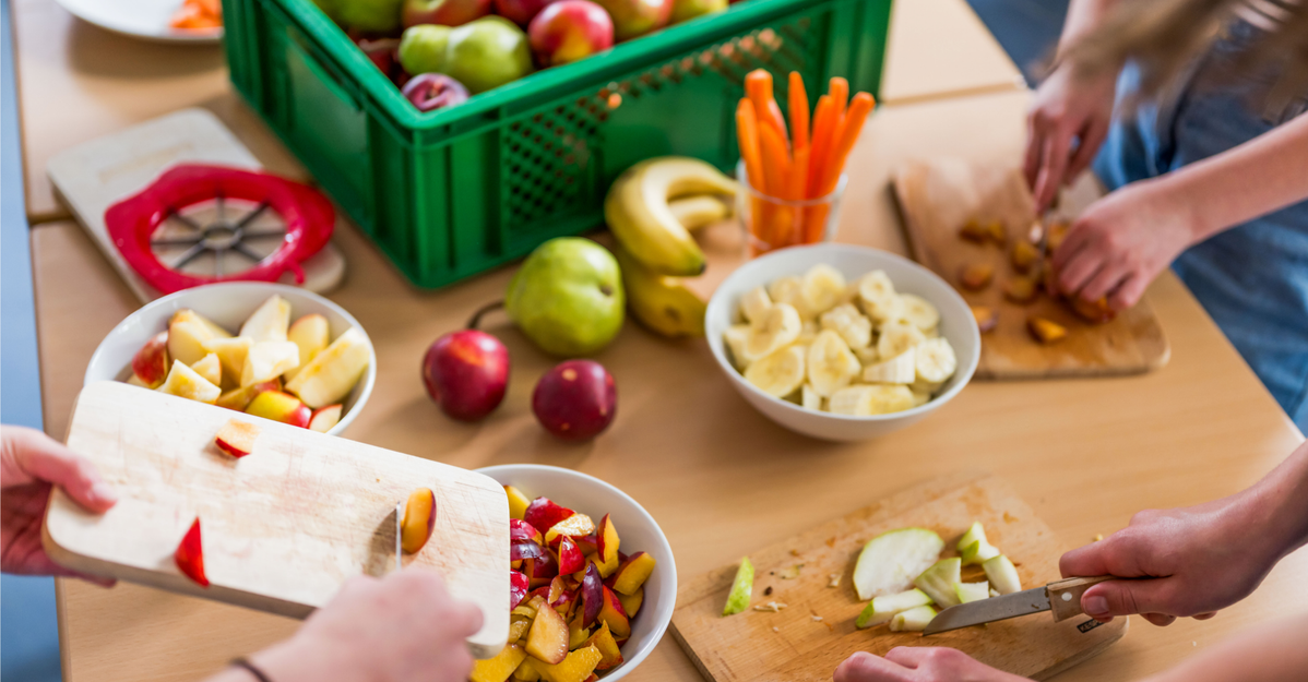 KI generiert: Menschen schneiden frisches Obst und Gemüse in einer Küche. Es gibt Äpfel, Bananen, Karotten und mehr.