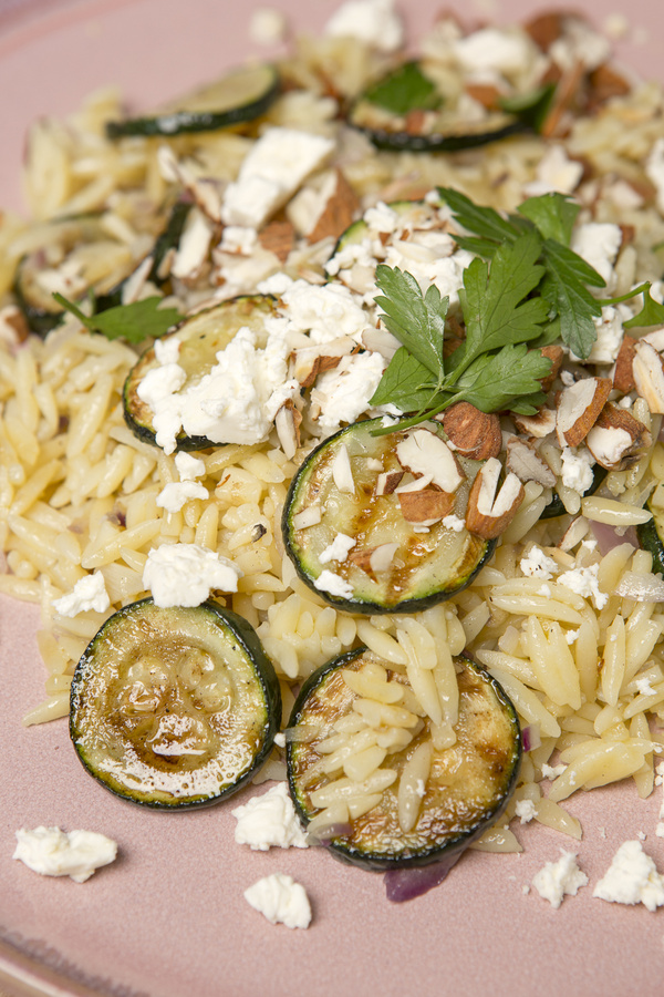 KI generiert: Nahaufnahme von Orzo-Nudeln mit Zucchini, Feta, Mandeln und Petersilie, auf einem rosa Teller serviert.