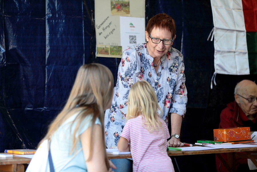 KI generiert: Eine ältere Frau spricht mit zwei Kindern an einem Tisch. Text im Hintergrund: "Gemeinsam den Hunger besiegen".