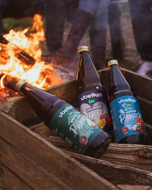 KI generiert: Das Bild zeigt drei Flaschen Voelkel Bio Punsch in einer Holzkiste vor einem brennenden Lagerfeuer. Die Flaschen sind in verschiedenen Geschmacksrichtungen etikettiert.
