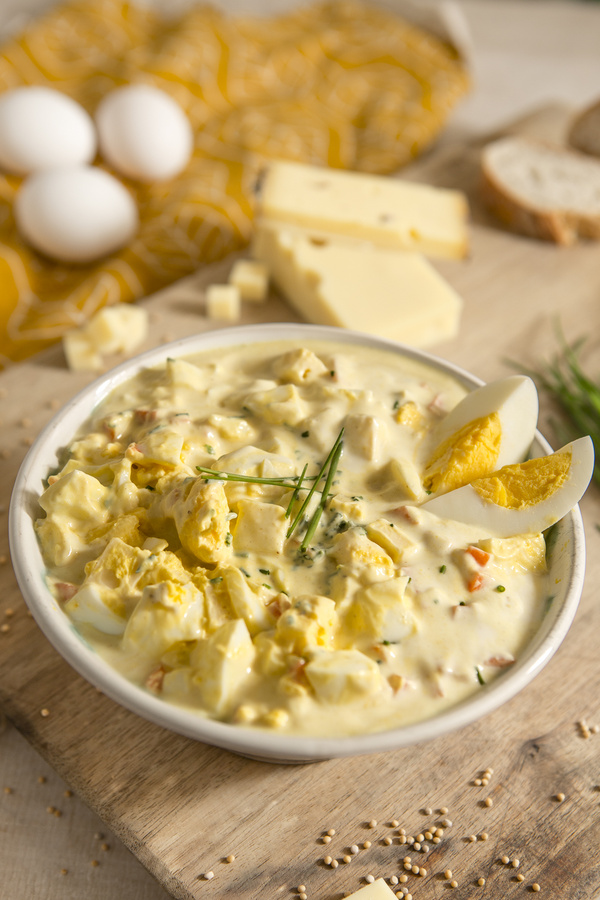 KI generiert: Ein Teller Eiersalat mit Kräutern, umgeben von Käse, Eiern und Brot auf einem Holztisch.