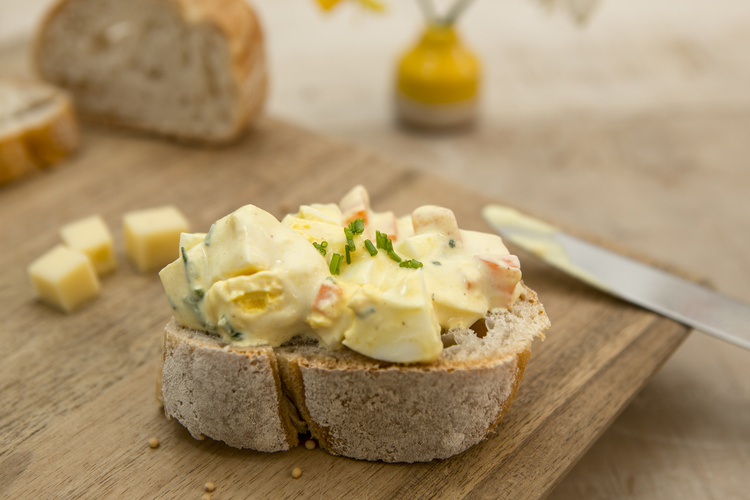 KI generiert: Ein Brotstück mit Eiersalat und Schnittlauch, im Hintergrund Käsewürfel.