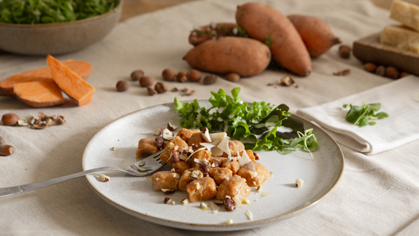 KI generiert: Ein Teller mit Gnocchi, Grünkohl und Nüssen, arrangiert auf einem Tisch mit Süßkartoffeln und Tuch.