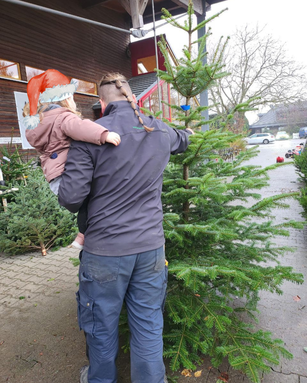 KI generiert: Ein Mann mit einem kleinen Kind auf dem Arm steht vor einem Weihnachtsbaum und scheint diesen zu inspizieren. Das Kind trägt eine rote Weihnachtsmütze.