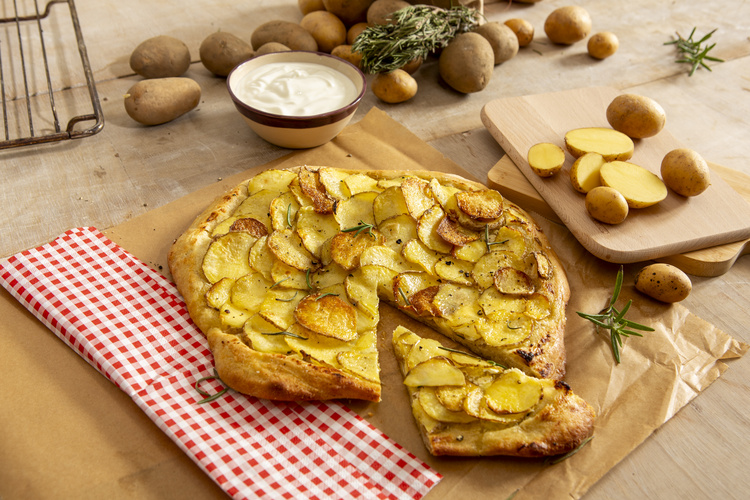 KI generiert: Kartoffelpizza mit einem Stück herausgeschnitten, umgeben von Kartoffeln und Kräutern auf Holzfläche.