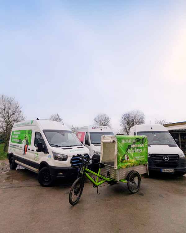 KI generiert: Drei Lieferwagen und ein Lastenfahrrad mit "frisch und grün - locker und echt - Bio live genießen" stehen nebeneinander.