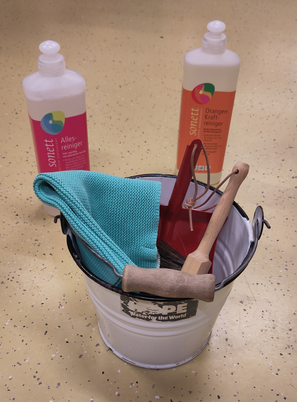 KI generiert: Ein Eimer mit Putzutensilien und zwei Flaschen Reinigungsmittel auf dem Boden. Text: "sonett Allesreiniger" und "Orangen Kraftreiniger".